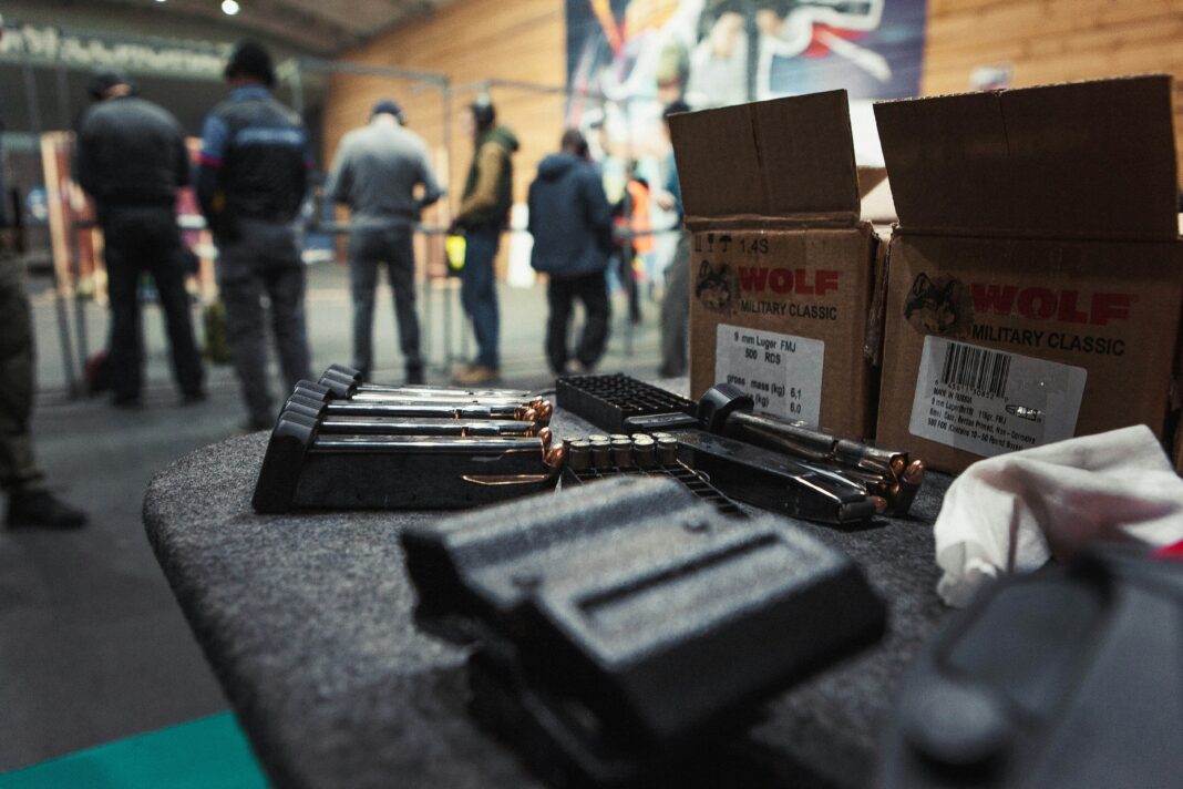 Boxes of ammunition and loaded magazines on a table at a shooting range with people standing in the background.