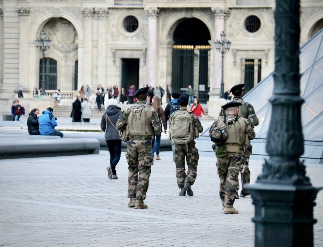 Soldiers in camouflage uniforms patrolling a public square near a historic building as civilians walk nearby.