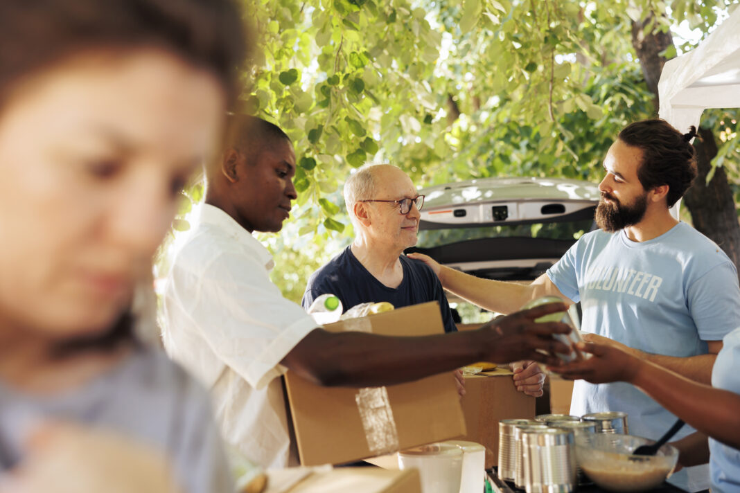 Volunteers helping a homeless man with food and supplies at a community outreach event under green trees.