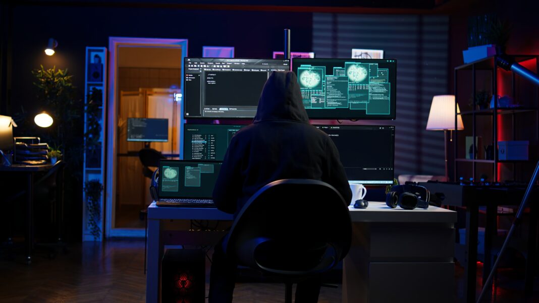 Hacker in dark room using multiple monitors to conduct cyberattack and steal sensitive data