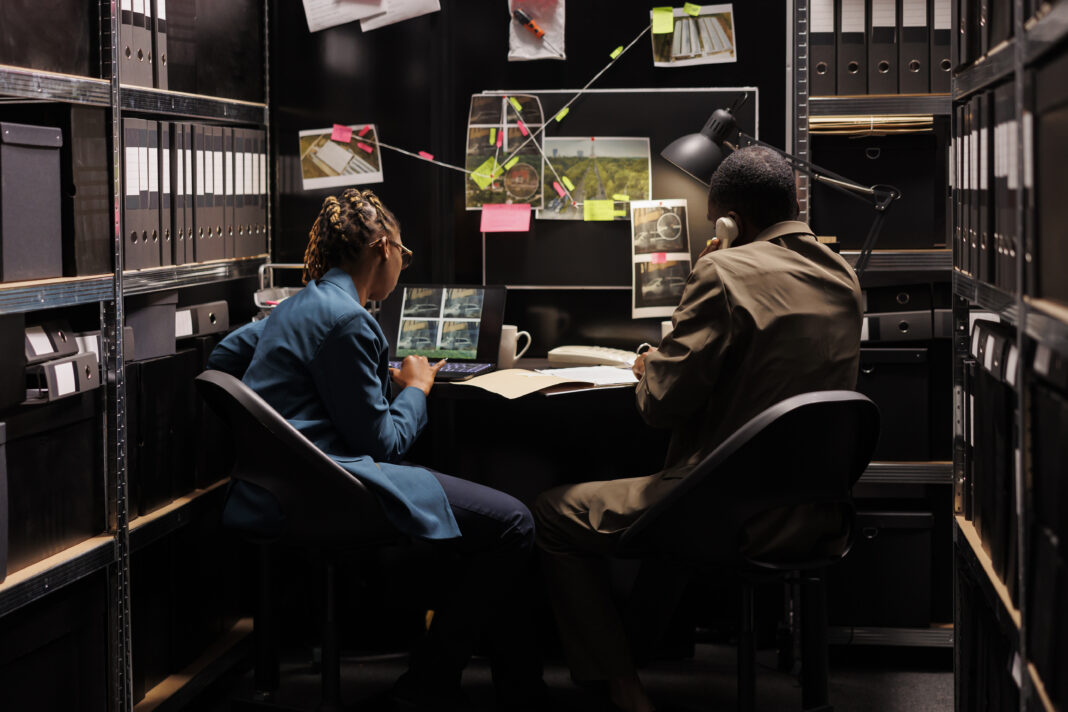 Two investigators sitting in an evidence room analyzing photos and case files on a board.