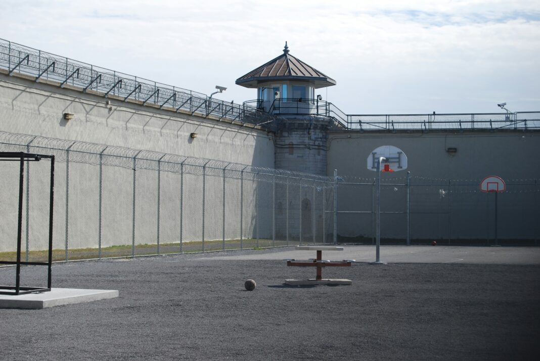 Empty prison yard with barbed-wire fences, a watchtower, and a basketball hoop.