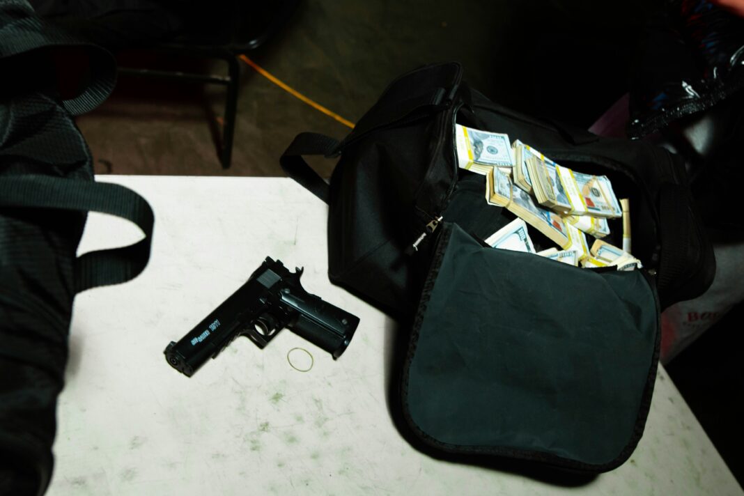 A handgun placed on a table beside a duffel bag filled with bundles of U.S. dollar bills.