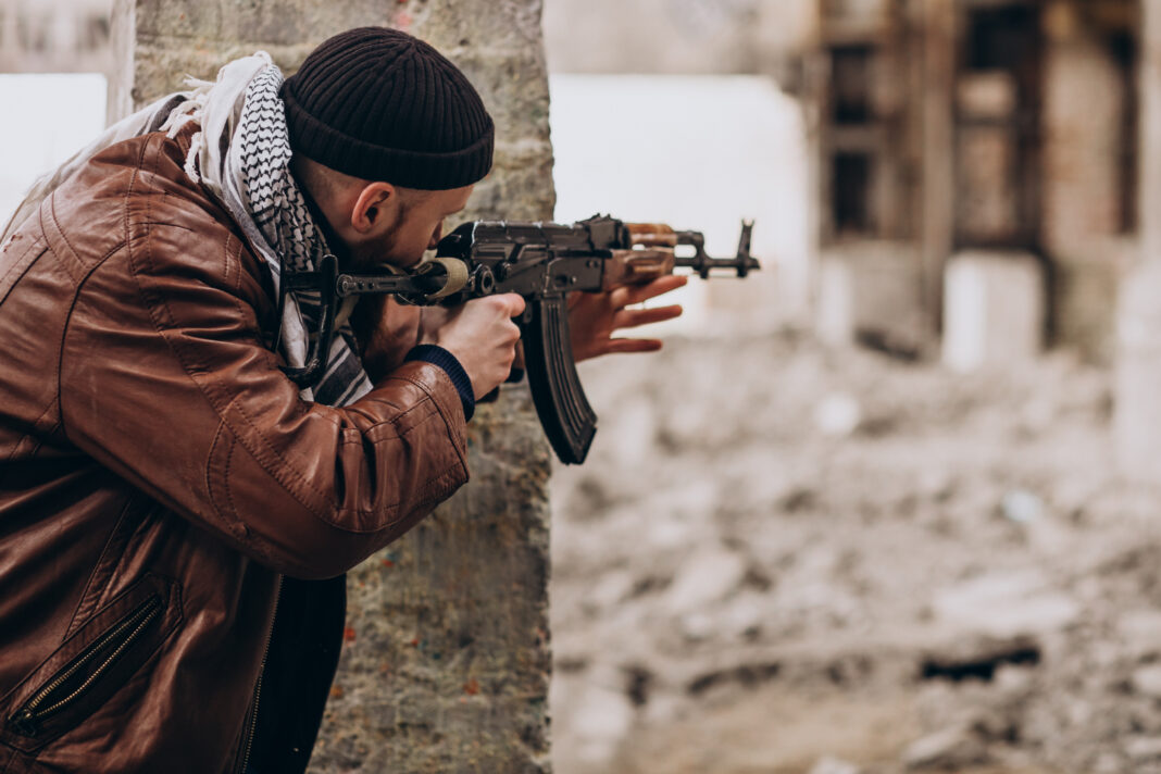 Armed man taking cover behind a wall and aiming a rifle in a conflict zone