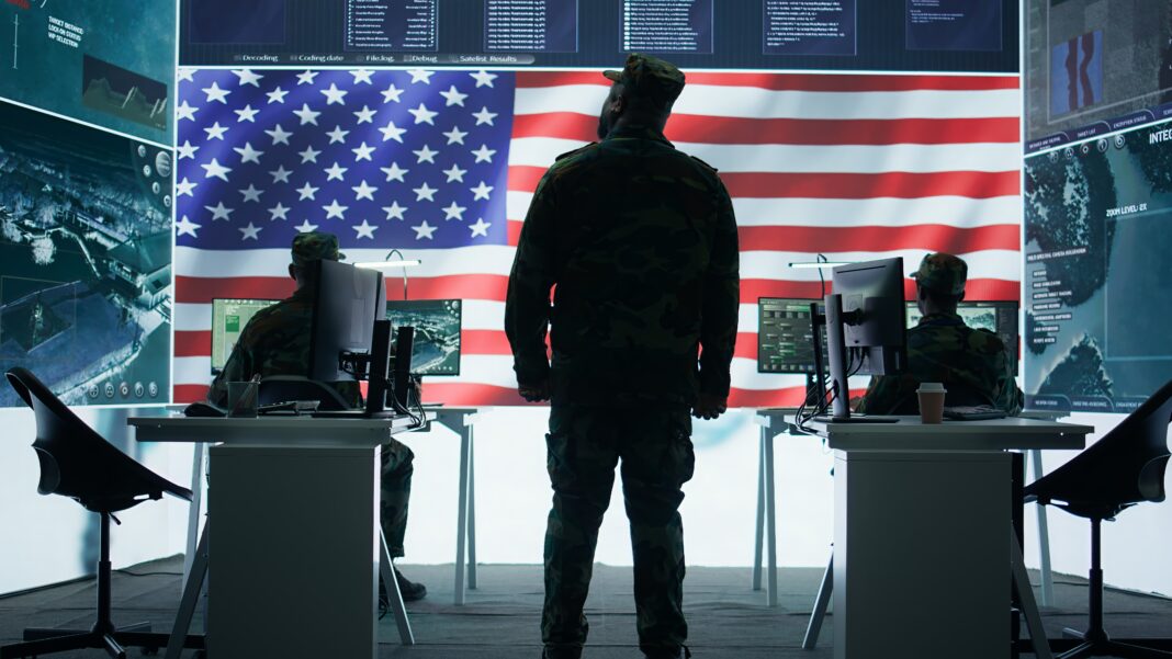 U.S. military officers in camouflage uniforms monitor computer systems with an American flag in the background.